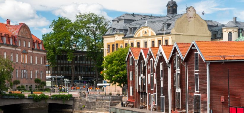 Gamla byggnader med röd taktegell och träd vid en kanal med framtida byggnader i bakgrunden.