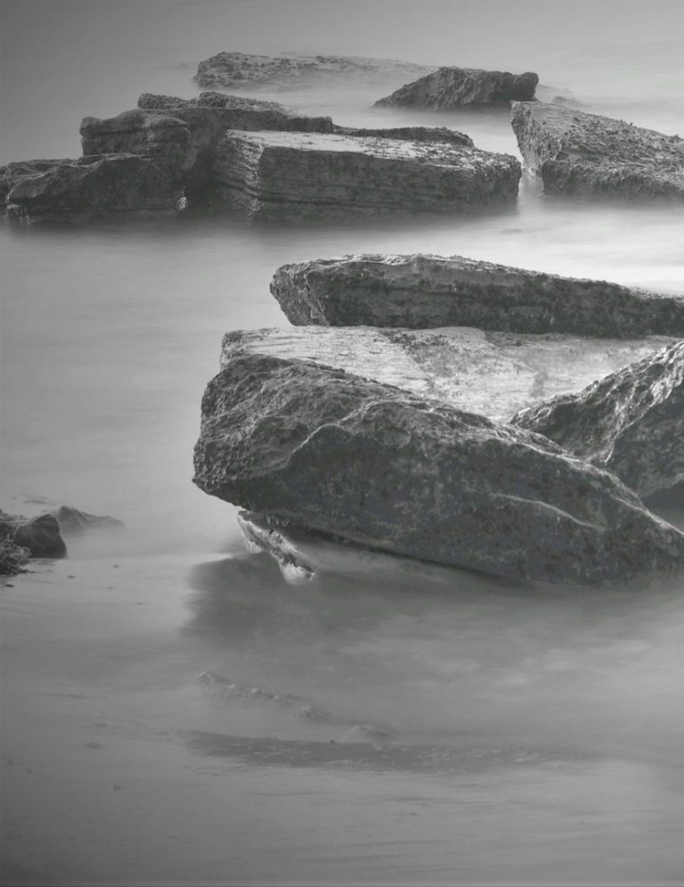 Grå stenar vid vatten, delvis täckta av mjukt, reflekterande vatten.