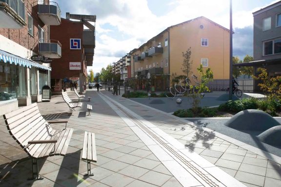 Gatumiljö med bänkar, träd och flerfamiljshus under en klar himmel.