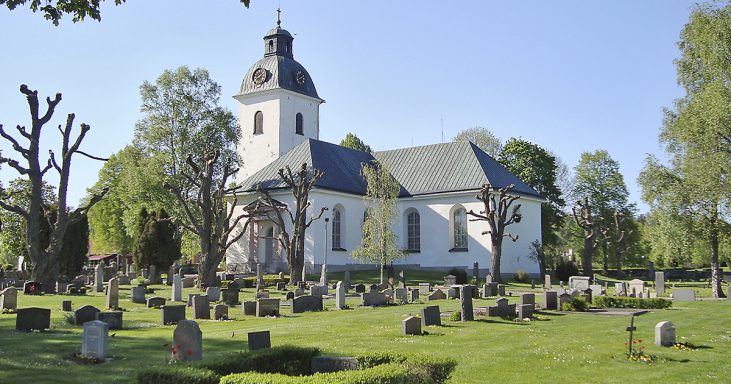 Vit kyrka med klocktorn omgiven av gröna träd och gravstenar i en lugn kyrkogård.